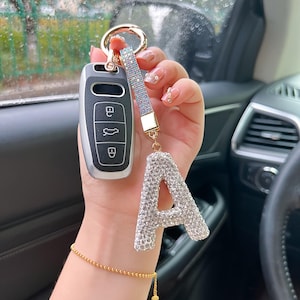 Könnte beinhalten: Ein Autoschlüsselanhänger mit einem silbernen und schwarzen Gehäuse, befestigt an einem glitzernden Riemen und einem großen, mit Strasssteinen besetzten Buchstaben "A"-Anhänger. Der Schlüsselanhänger hat einen goldfarbenen Ring.