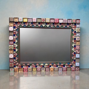 May include: Rectangular decorative mirror with a mosaic tile frame. The frame features colorful square tiles with various patterns and colors, including red, blue, yellow, and green. The mirror has a brown inner frame and a reflective surface.