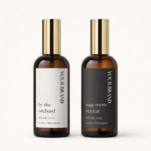 Op de afbeelding: Twee bruine glazen spuitflessen met goudkleurige doppen. Eén etiket leest "by the orchard" en de andere "sage moss retreat". Elke fles bevat 100 ml room-linnenspray. De merknaam staat verticaal afgedrukt.