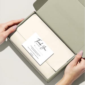Puede incluir: Una caja verde claro abierta, que revela un interior blanco con un artículo doblado y una tarjeta blanca. La tarjeta presenta una escritura negra que dice "Thank You" y texto adicional. La caja está siendo abierta.