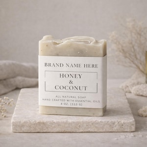 May include: A bar of Honey & Coconut soap, hand-crafted with essential oils, sits on a stone block. The soap is wrapped with a label that reads "BRAND NAME HERE" and "HONEY & COCONUT". The soap weighs 4 oz. A bottle of oil and decorative elements are in the background.