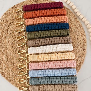 May include: A collection of macrame keychains in various colors, including burgundy, orange, and blue. Each keychain features a woven design and a gold-colored clasp. The keychains are arranged on a woven placemat.