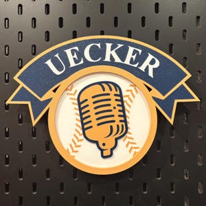 Puede incluir: Un letrero de madera con el nombre "UECKER" en blanco sobre una pancarta azul marino. El letrero tiene un tema de béisbol, con un diseño de micrófono naranja y un diseño de costura de béisbol. Montado en una placa perforada negra.