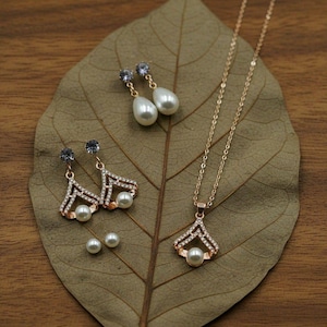 Puede incluir: Un conjunto de joyas con un collar y pendientes dorados con perlas de imitación y cristales transparentes. El conjunto incluye un collar con colgante, dos pares de pendientes y dos pequeños pendientes de perlas. Las joyas se exhiben sobre una hoja grande y seca.