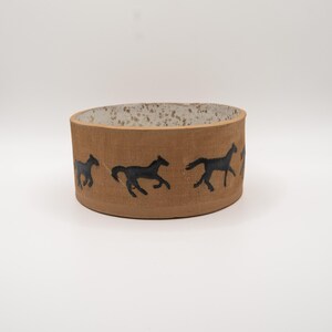 Può includere: Una ciotola cilindrica in ceramica con un esterno color marrone chiaro caratterizzato da un motivo ripetitivo di cavalli neri al galoppo. L'interno della ciotola presenta una base bianca con macchie marroni, creando un netto contrasto.