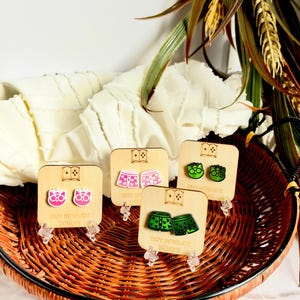 May include: Four pairs of stud earrings displayed on individual square wooden cards. The earrings feature designs of pink cat faces with glasses, pink boxer shorts with hearts, green cat faces with glasses, and green boxer shorts with hearts.