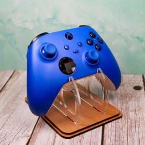 May include: A blue Xbox controller rests on a wooden and clear acrylic stand. The controller has black buttons and joysticks. The stand is made of wood and clear acrylic, designed to hold the controller upright.