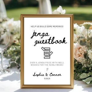 Könnte beinhalten: Ein gerahmtes Schild mit goldenem Rahmen, das die Worte "jenga guestbook" in Schreibschrift zeigt. Das Schild enthält den Text "Help us build some memories" und "Sign a Jenga piece with well wishes for the newlyweds!" mit den Namen Sophia & Connor und dem Datum 10.06.25.
