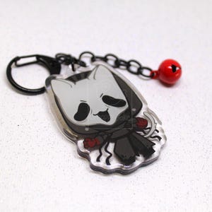 May include: An acrylic keychain featuring a cartoon character with a white cat-like mask, black hooded cloak, and red accents. The charm is attached to a black metal lobster clasp and chain with a small, round, red jingle bell attached.