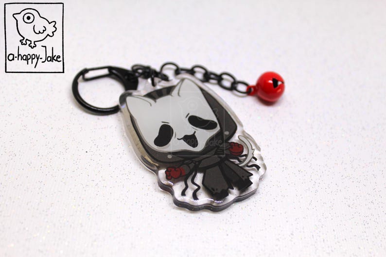 May include: An acrylic keychain featuring a cartoon character with a white cat-like mask, black hooded cloak, and red accents. The charm is attached to a black metal lobster clasp and chain with a small, round, red jingle bell attached.