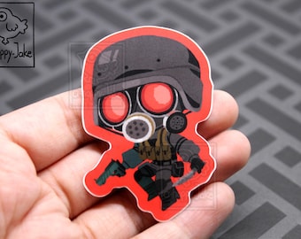 May include: A die-cut sticker featuring a chibi-style character wearing a dark tactical helmet, a gas mask with glowing red circular lenses, and dark combat gear. The character holds a small handgun and a knife against a bright red background.