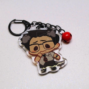 May include: An acrylic keychain featuring a cartoon character with dark hair, glasses, a white shirt, and a striped tie. The charm is attached to a black metal ring and chain with a small red bell. The character has a worried expression.