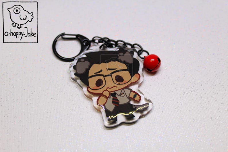 May include: An acrylic keychain featuring a cartoon character with dark hair, glasses, a white shirt, and a striped tie. The charm is attached to a black metal ring and chain with a small red bell. The character has a worried expression.