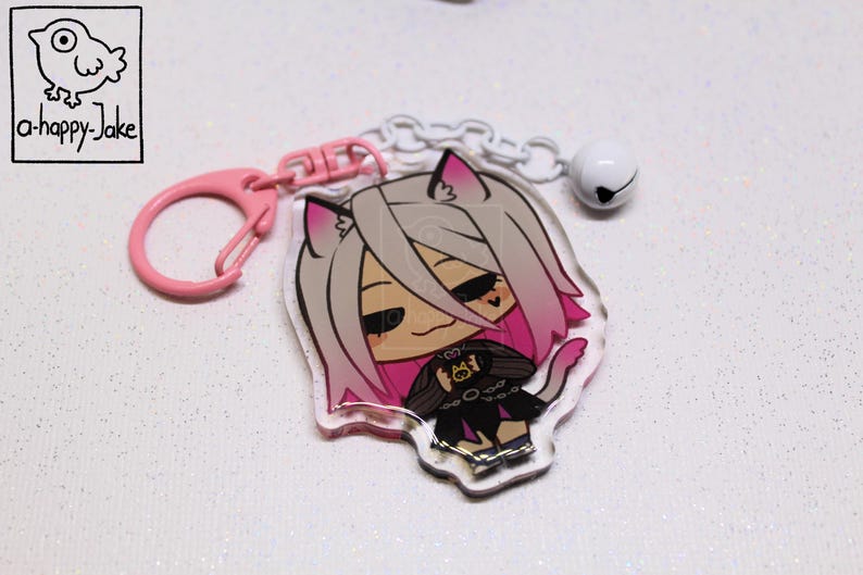 May include: An acrylic keychain featuring a cartoon character with light gray and pink hair, cat ears, and a black outfit. The charm is attached to a pink plastic clasp and a white chain with a small white bell. The logo a-happy-Jake is visible.