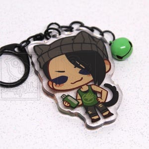May include: An acrylic keychain featuring a cartoon character with black hair, a gray beanie with cat ears, and a green tank top holding a green spray paint can. The keychain includes a black metal ring and a small green bell charm.