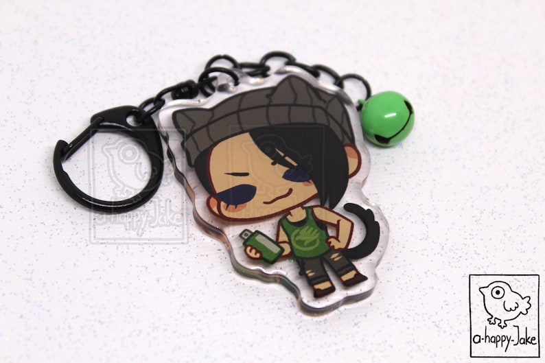May include: An acrylic keychain featuring a cartoon character with black hair, a gray beanie with cat ears, and a green tank top holding a green spray paint can. The keychain includes a black metal ring and a small green bell charm.