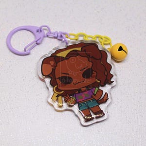 May include: An acrylic keychain featuring a cartoon character with brown hair and dog ears, wearing a purple crop top and blue shorts. The charm is attached to a light purple plastic clasp and a yellow chain with a small yellow bell attached.