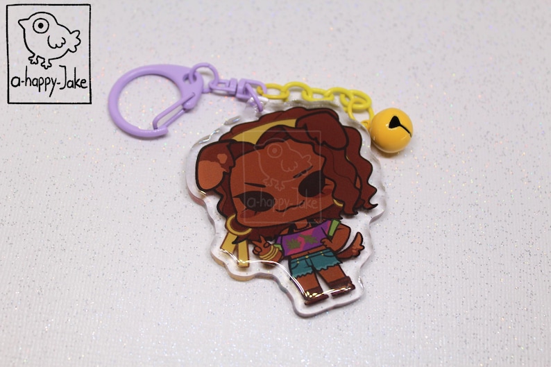 May include: An acrylic keychain featuring a cartoon character with brown hair and dog ears, wearing a purple crop top and blue shorts. The charm is attached to a light purple plastic clasp and a yellow chain with a small yellow bell attached.