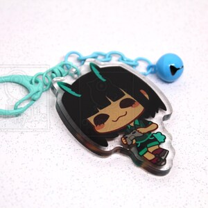 May include: An acrylic keychain featuring a cartoon character with black hair and teal horns, holding a gray game controller. The charm is attached to a light blue plastic chain with a matching light blue bell. The text a-happy-jake is visible.