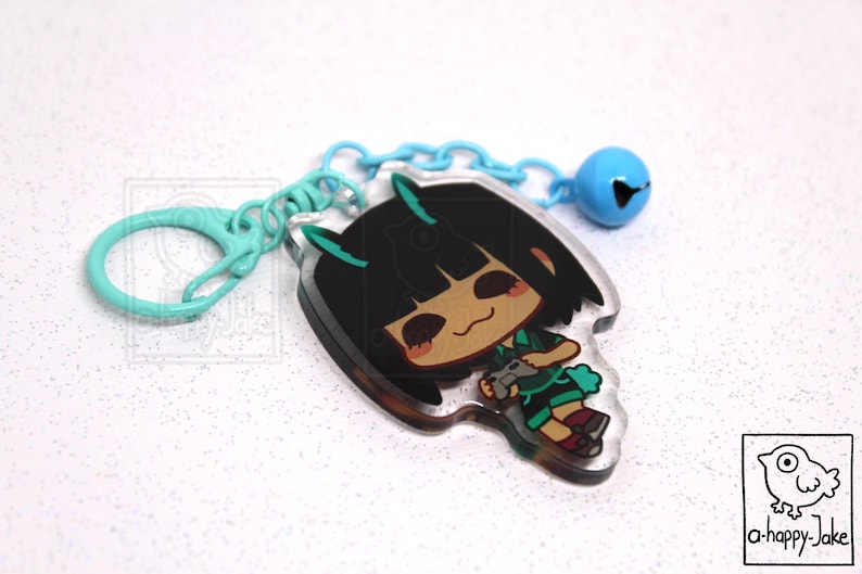 May include: An acrylic keychain featuring a cartoon character with black hair and teal horns, holding a gray game controller. The charm is attached to a light blue plastic chain with a matching light blue bell. The text a-happy-jake is visible.