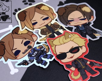 May include: Four die-cut vinyl stickers featuring chibi-style characters with animal ears and tails. One character wears an R.P.D. uniform, another a brown jacket, one a dark coat, and one a black outfit with sunglasses. A logo with a bird reads a-happy-jake.
