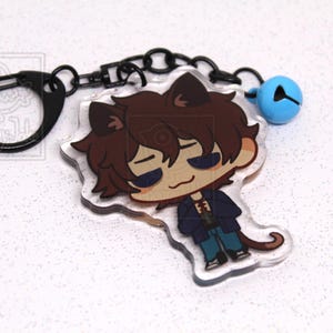 May include: An acrylic keychain featuring a cartoon character with brown hair and cat ears, wearing a dark blue jacket and blue pants. The charm is attached to a black metal ring and chain with a small light blue bell. A logo reads a-happy-jake.