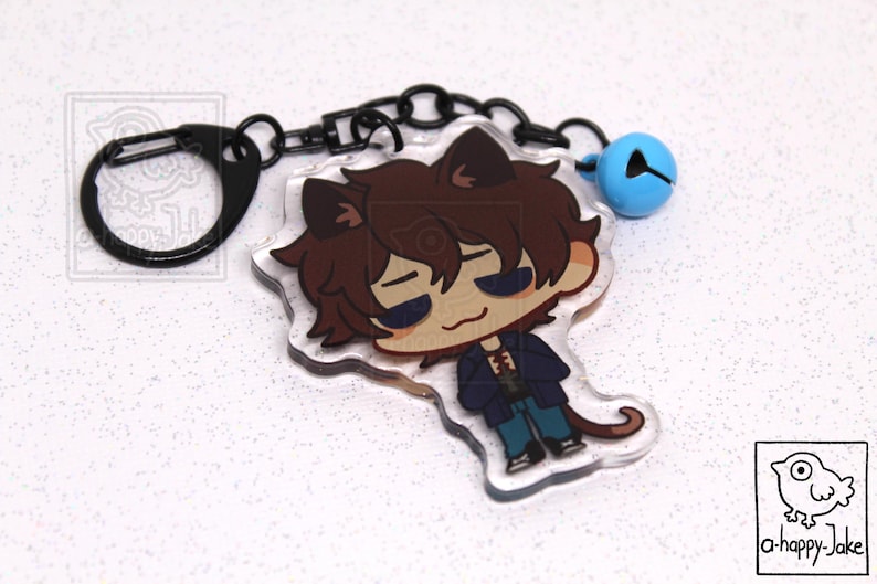 May include: An acrylic keychain featuring a cartoon character with brown hair and cat ears, wearing a dark blue jacket and blue pants. The charm is attached to a black metal ring and chain with a small light blue bell. A logo reads a-happy-jake.