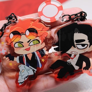 May include: A double-sided acrylic keychain featuring two cartoon characters. One character has orange hair and sunglasses, the other has black hair and a suit. The keychain includes a black chain and two small red and white life preserver charms.