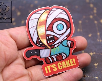 May include: A die-cut sticker featuring a cartoon mummy character with one side of its head sliced open to reveal yellow cake layers. The mummy holds a silver kitchen knife and wears a teal shirt. The text IT'S CAKE! is printed in yellow.