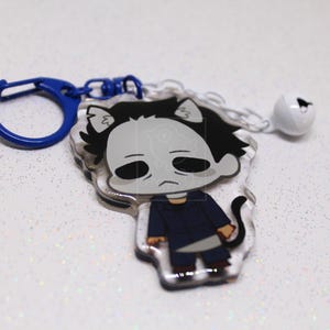 May include: An acrylic keychain featuring a cartoon character with a white mask, black hair, and a dark blue jumpsuit, holding a silver knife. The charm has cat ears and a tail, attached to a blue plastic clasp and a small white jingle bell.