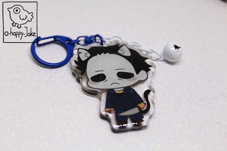 May include: An acrylic keychain featuring a cartoon character with a white mask, black hair, and a dark blue jumpsuit, holding a silver knife. The charm has cat ears and a tail, attached to a blue plastic clasp and a small white jingle bell.