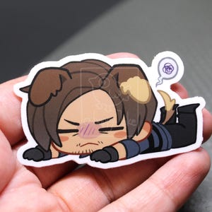 May include: A die-cut vinyl sticker featuring a chibi-style character with brown dog ears, dark brown hair, and a stubble beard, lying on their stomach. The character wears a dark blue shirt and black pants, with a purple scribble thought bubble.