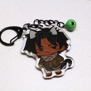 May include: An acrylic keychain featuring a cartoon character with dark hair, cat ears, and a brown jacket, attached to a black metal ring and a small green bell. The top left corner displays a square logo with a bird and the text a-happy-Jake.