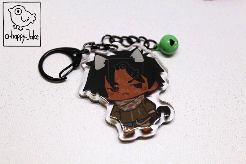 May include: An acrylic keychain featuring a cartoon character with dark hair, cat ears, and a brown jacket, attached to a black metal ring and a small green bell. The top left corner displays a square logo with a bird and the text a-happy-Jake.