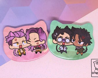 May include: Two cat-shaped pinback buttons featuring anime-style characters. The pink button shows two characters with purple hair and cat ears. The green button shows two characters with dark hair and cat ears. A small logo with a bird and the text a-happy-jake is visible.
