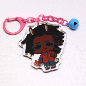 May include: An acrylic keychain featuring a cartoon character with dark hair, animal ears, and blue glasses, wearing a red shirt and blue pants. The charm is attached to a pink plastic ring and chain with a small blue bell.