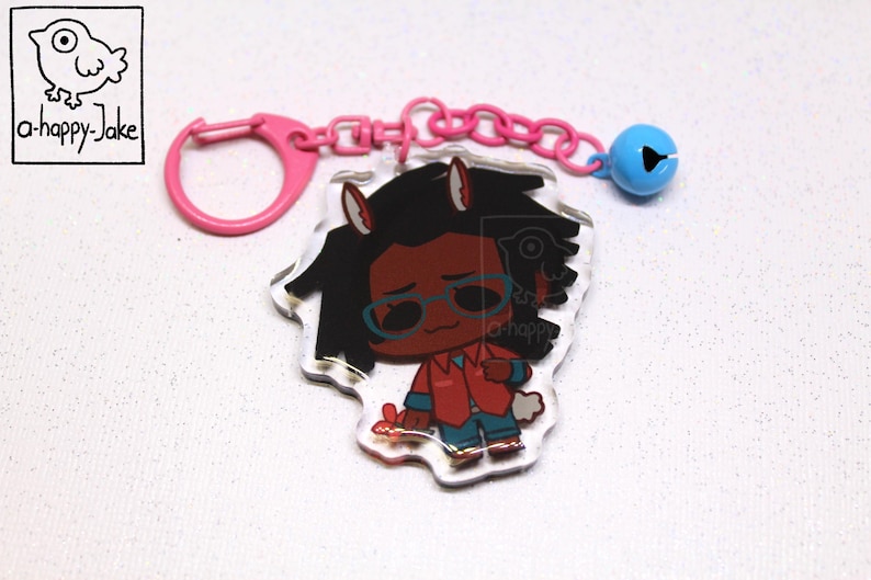 May include: An acrylic keychain featuring a cartoon character with dark hair, animal ears, and blue glasses, wearing a red shirt and blue pants. The charm is attached to a pink plastic ring and chain with a small blue bell.