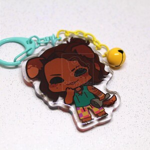May include: An acrylic keychain featuring a cartoon character with brown fur, wearing a green shirt and checkered pants, holding a gray plunger. The charm is attached to a light blue plastic clasp and a yellow chain with a small yellow bell.