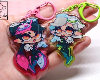 May include: Two acrylic keychains featuring stylized cartoon characters with pointed ears and fangs. One character has dark hair with pink accents and a pink clasp, while the other has light hair with green accents and a green clasp. Both display a logo.