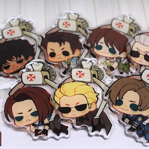 May include: A collection of seven clear acrylic charms featuring chibi-style characters from Resident Evil, each being lifted by a claw machine mechanism with an Umbrella Corporation logo. The charms depict various characters with distinct hair colours.