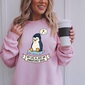 May include: A light pink sweatshirt featuring a cartoon penguin wearing a blue beanie and sitting on an ice floe. The penguin is in a thought bubble with a lit match. The banner below reads "MY LIFE IS JUST AN UNMATCHED SOCK."