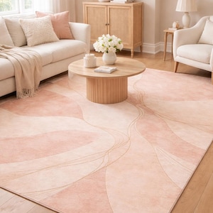 Può includere: Un grande tappeto rettangolare con un design morbido e astratto nei toni del rosa e del crema. Il tappeto presenta linee curve e fluide in un colore oro più chiaro. Il tappeto è collocato in un soggiorno con un divano, sedie e un tavolino di legno.