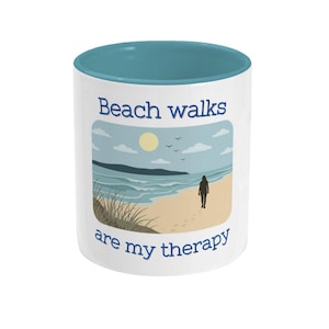 May include: White ceramic mug with a blue interior. The mug features a beach scene with the text "Beach walks are my therapy". The design includes a person walking on the beach, the sun, clouds, and ocean waves.