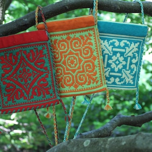 May include: Three rectangular crossbody bags with braided straps and tassels, featuring intricate geometric patterns. The first bag is red with teal details, the second is orange with light green details, and the third is teal with white details.