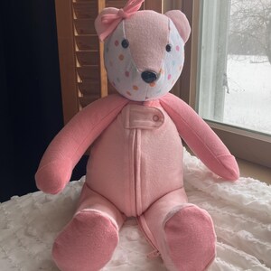May include: A handmade pink teddy bear with a bow on its head. The bear's face is white with colorful polka dots. It has a black nose and is wearing a pink onesie with a zipper. The bear is sitting on a white, textured surface.