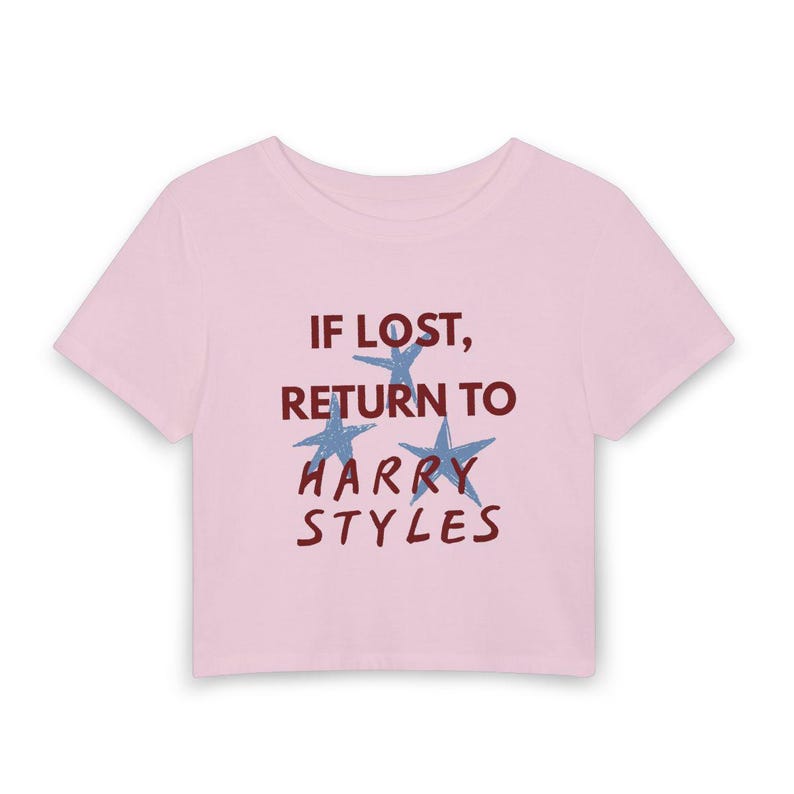 May include: A light pink short-sleeved crop top featuring the dark red text "IF LOST, RETURN TO HARRY STYLES" printed on the front. The design includes three light blue star graphics interspersed between the lines of text on the center chest.