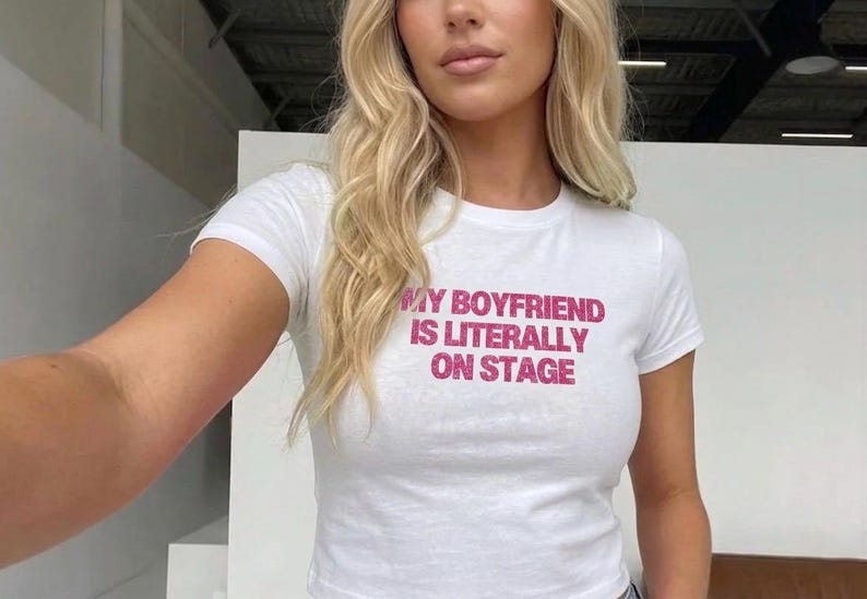 May include: White, short-sleeved t-shirt with the text "MY BOYFRIEND IS LITERALLY ON STAGE" in pink. The shirt is a fitted style. The model has blonde hair.