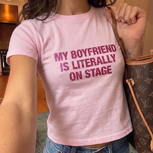 May include: A light pink t-shirt with the text "MY BOYFRIEND IS LITERALLY ON STAGE" in a glittery, magenta font. The shirt is paired with blue jeans. A brown and tan handbag is visible in the image.