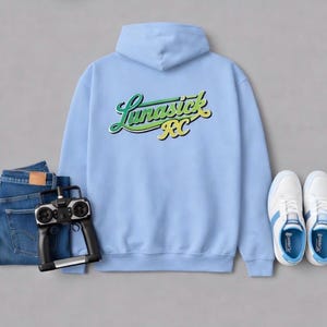May include: A light blue hoodie with the text "Lunasick RC" in green and yellow script. The image also includes a pair of blue jeans, a black remote control, and white sneakers with blue accents.