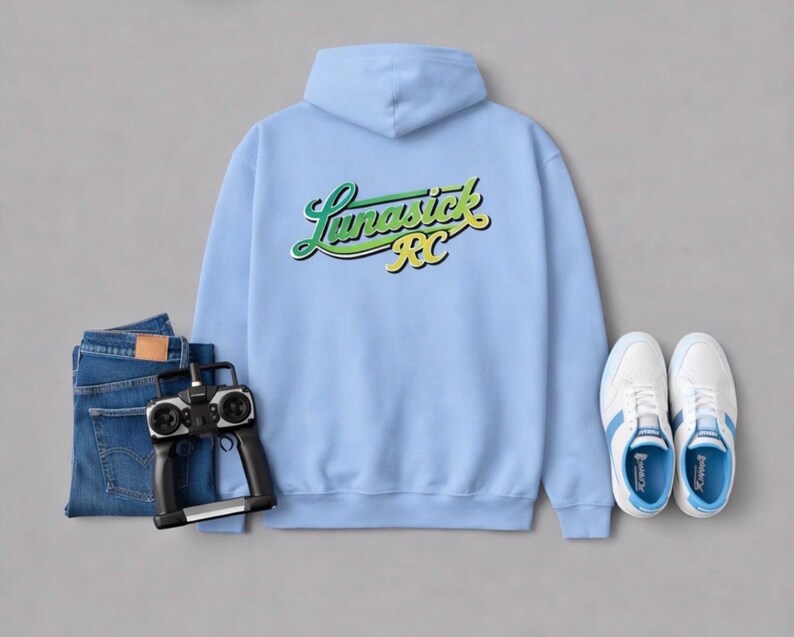 May include: A light blue hoodie with the text "Lunasick RC" in green and yellow script. The image also includes a pair of blue jeans, a black remote control, and white sneakers with blue accents.